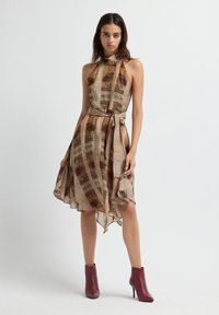 Vrouw in een mouwloze, hooggesloten bruine gestreepte jurk met een asymmetrische zoom en riem, gecombineerd met rode enkellaarsjes, staande tegen een witte achtergrond.