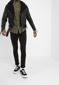 Chaqueta de cuero negra estilo motorista con cuello de borrego, camisa de botones color oliva, pantalones ajustados negros y zapatillas negras con suelas blancas.