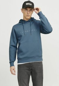 Sininen huppari, jossa on etu-kangaroo-tasku, narut ja valkoinen teksti "JACK & JONES". Yhdistetty harmaisiin farkkuihin ja mustaan lippikseen.