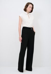 Weiße ärmellose Bluse mit einem Schlüssel-ausschnitt, kombiniert mit hochtaillierten schwarzen Hosen. Verfügt über einen schwarzen Gürtel und ein minimalistisches goldfarbenes Armband.
