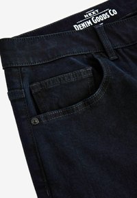 Donkerdenim jeans met een gladde textuur, met een achterzak en een klein knoopschema. Etiket zegt "NEXT DENIM GOODS CO."