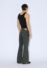 Osoba plecami do widza, ubrana w czarny top na ramiączkach i ciemnoszare dżinsy z wysokim stanem i rozszerzanymi nogawkami, z beżową metką na pasku.