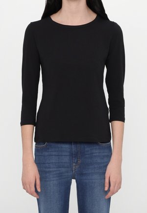 Femme portant un t-shirt noir uni à manches trois-quarts et un jean bleu, montrée du cou jusqu'aux hanches sur fond blanc.