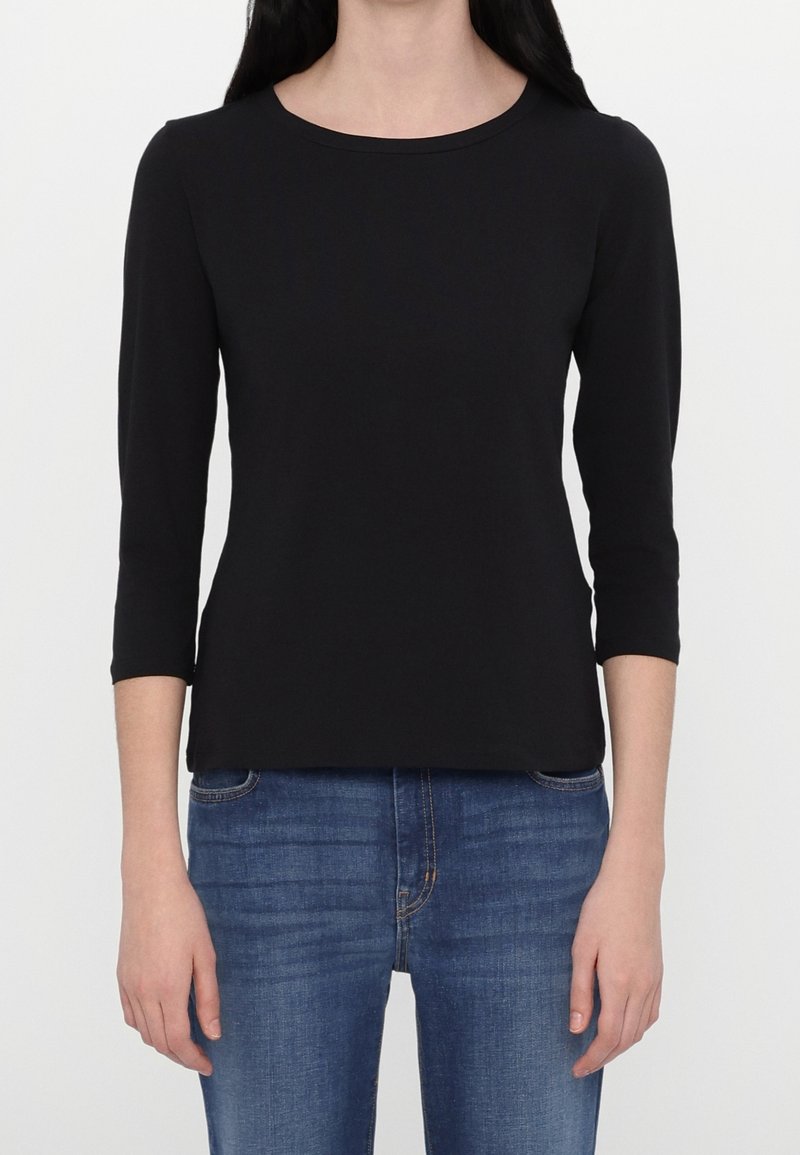 Femme portant un t-shirt noir uni à manches trois-quarts et un jean bleu, montrée du cou jusqu'aux hanches sur fond blanc.