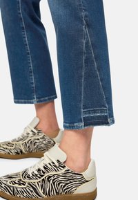 Sneaker mit Zebramuster in Schwarz und Weiß, cremefarbenem Absatz, beigen Akzenten, Schnürsenkeln und brauner Sohle; kombiniert mit blauen, gekürzten Jeans mit ausgefranstem Saum.