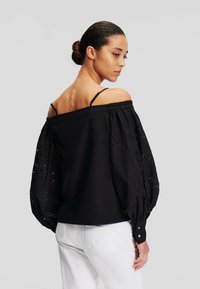 Top nero scollato con spalle scoperte, maniche in pizzo a sbuffo, con dettagli ricamati e una leggera lucentezza. Abbinato a jeans bianchi.