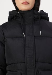 Svart pufferjacka med hög krage och justerbara snoddar, med en slät, något skrynklig yta och sömdetaljer.