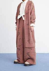 Rosa texturerad stickad cardigan med Nike-logomönster, lager på lager med matchande byxor, i kombination med kraftiga ljusa skor.