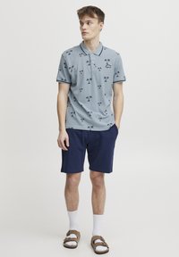 Korteermet, lyseblå poloshirt med svarte palme mønstre, parret med marineblå shorts. Brukt med hvite sokker og brune sandaler.