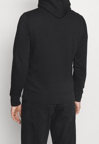Sudadera negra con mangas largas, capucha con cordón y dobladillo acanalado. El material parece suave, con una textura lisa y un minimalismo en los detalles.