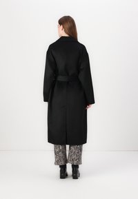 Abrigo largo negro con cinturón, que presenta un cuello de pico y corte recto. Llevado sobre pantalones holgados con estampado y botas negras gruesas.