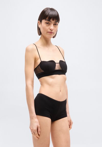 Γυναίκα με κοντά σκούρα μαλλιά, φορούσα μαύρο μπραλέτο σε σχήμα καρδιάς και μαύρο σορτς με ψηλή μέση, μπροστά σε απλό λευκό φόντο.