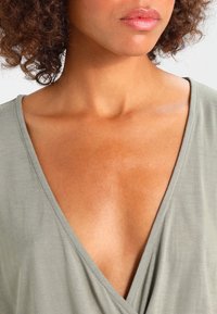 Un haut en tissu léger olive, avec un décolleté en V profond et une coupe décontractée, mettant en valeur la texture lisse de la peau autour du cou.