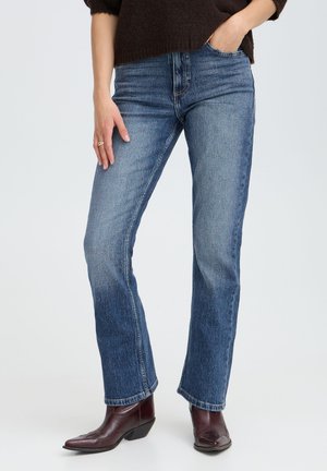Personne portant un jean bleu droit, des bottines en cuir marron et un pull marron foncé, une main dans la poche et l'autre détendue sur le côté.