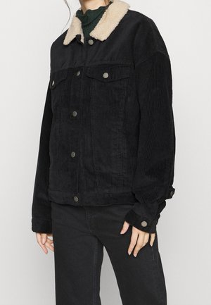 Personne portant une veste en velours côtelé noire avec des boutons argentés et un col en polaire beige, associée à un pantalon noir et un haut vert foncé.