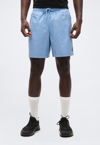 Άνδρας που φορά ανοιχτό μπλε σορτς με κορδόνι, λευκές κάλτσες και μαύρα αθλητικά παπούτσια, στέκεται μπροστά σε απλό φωτεινό φόντο.