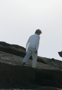 Witte lange mouwen shirt en lichtgekleurde broek. Persoon loopt blootsvoets over een rotsachtig terrein tegen een heldere, overbelichte achtergrond.