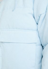 Helle blaue Cord-Puffjacke mit strukturierter Oberfläche, ausgestattet mit einer vorderen Pattaschen und einem Reißverschluss am Kragen.