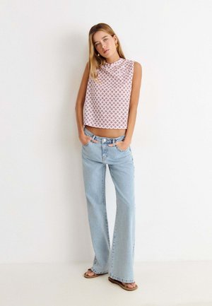Jeune femme aux longs cheveux blonds portant un haut sans manches à motifs, un jean bleu clair à jambes larges et des sandales, se tient contre un mur blanc.