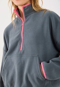 Dark gray fleece pullover with a pink zip and neck lining. Features a front pocket and contrasting cuffs, emphasizing comfort and warmth.