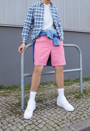 Osoba ubrana w różowe szorty, białe trampki, białe skarpetki, biały t-shirt, niebieską koszulę w kratę oraz niebieski sweter zawiązany wokół talii, stojąca przy metalowej poręczy.