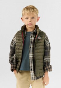 Ragazzo biondo che indossa un gilet trapuntato verde sopra una camicia a quadri con le maniche arrotolate e pantaloni beige, in piedi con le mani nelle tasche.