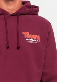 Mand iført en mørkerød hoodie med "Tommy JEANS, N.Y."-logo og tekst på venstre brystområde. Hoodieens snore er synlige.