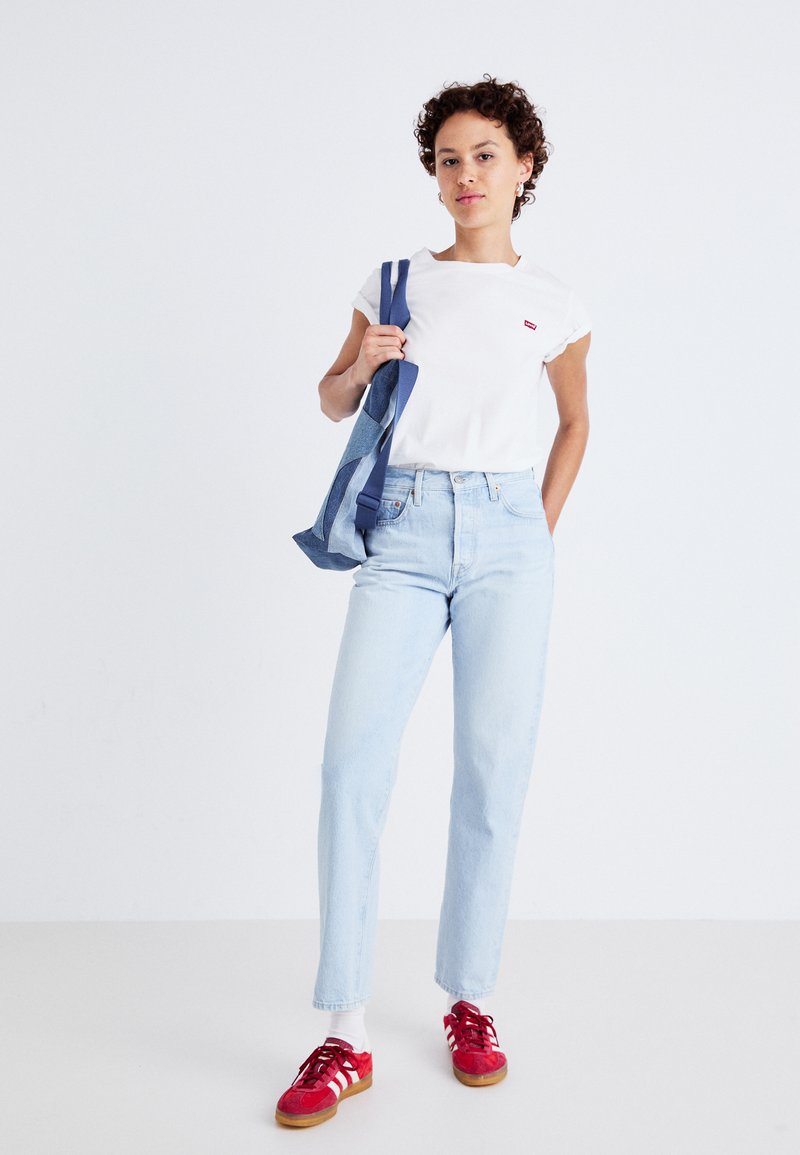 Maglietta bianca con maniche arrotolate e piccolo logo rosso, jeans a vita alta blu chiaro, scarpe da ginnastica rosse con dettagli bianchi, che tiene una borsa blu.