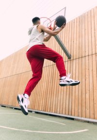 A person in a gray sleeveless hoodie and red joggers jumps toward a basketball hoop while holding a black basketball. The background is wooden.