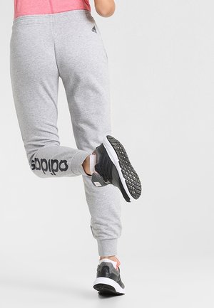 Person iført grå Adidas joggingbukser og sorte løbesko, der løfter ét ben bagud mod en ensfarvet lys baggrund.
