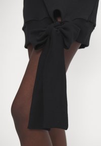 Sudadera negra con un detalle de lazo anudado en el dobladillo, con un corte en la parte superior y textura acanalada. La tela suave complementa un ajuste atlético.