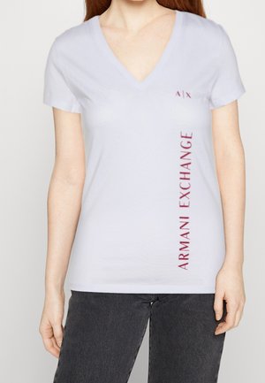 Vrouw draagt een witte V-hals T-shirt met korte mouwen en verticale rode tekst "ARMANI EXCHANGE" aan de rechterkant.