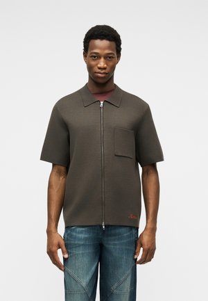 Jeune homme portant une chemise zippée à manches courtes, gris foncé, avec une poche poitrine, et un jean bleu, debout devant un fond uni.