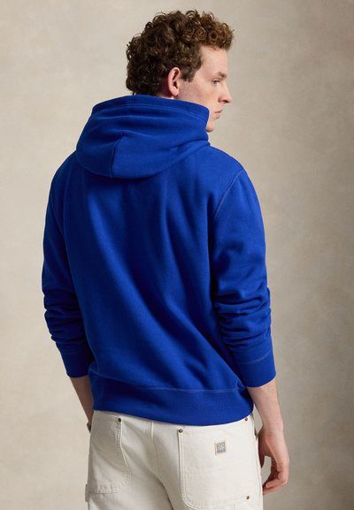 Homme aux cheveux bouclés portant un sweat à capuche bleu vif et un pantalon blanc, de profil sur un fond beige uni.