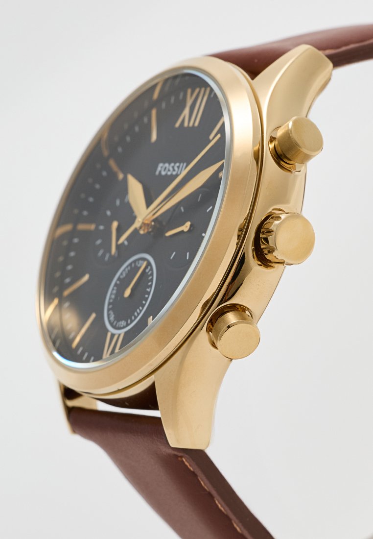 Reloj analógico en tono dorado con esfera negra, correa de cuero marrón y tres botones laterales para funcionalidad. Presenta números romanos y un sub-dial.