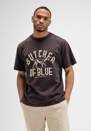 Man met kort haar en baard, die een zwart T-shirt met een hond en de tekst "Butcher of Blue" draagt, en beige broek, staand met handen in de zakken.