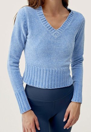 Femme portant un pull en maille côtelée bleu clair à col en V et un pantalon bleu foncé ajusté, debout, les mains détendues près des hanches.