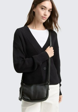 Borsa a tracolla in pelle nera texture, con una forma arrotondata, dotata di una tracolla e un logo discreto. Indossata con un cardigan nero e una maglietta bianca.