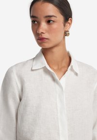Camicia bianca in lino con colletto classico, chiusura a bottoni e tessuto testurizzato. Il modello indossa orecchini a cerchio dorati. Design semplice ed elegante.