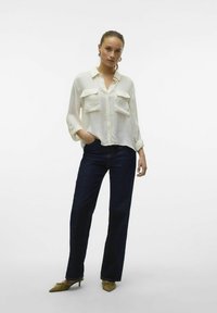 Camicia bianca leggera con bottoni e due tasche sul petto, abbinata a jeans a gamba larga blu scuro e scarpe verde oliva a punta.