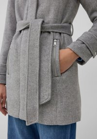 Manteau en mélange de laine gris avec une taille ceinturée, présentant une finition texturée et des poches zippées sur les côtés. Les poignets ont des fermetures à pression, complétés par du denim bleu.