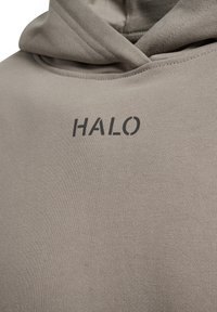 Nærbillede af beige hættetrøje stof med ordet "HALO" trykt med sort tekst nær halsen og hætten hvilende ovenpå.