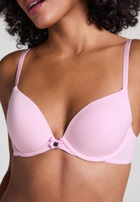 Reggiseno imbottito rosa chiaro con tessuto morbido, spalline regolabili e un dettaglio decorativo con fiocco e accessori al centro.