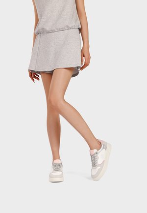 Donna che indossa un vestito casual grigio e sneakers grigie, bianche e rosa, in piedi con una gamba incrociata sull'altra su uno sfondo bianco.
