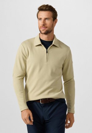 Man draagt een beige trui met lange mouwen en een kwart ritssluiting over een zwart overhemd en donkerblauwe broek met een bruine riem, kijkt iets naar rechts, neutrale achtergrond.