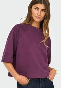 Femme aux cheveux longs châtain clair portant une chemise violette ample à manches courtes et un jean foncé, les mains dans les poches, regardant légèrement sur le côté.