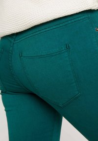 Jeans verde azulado con un ajuste ceñido, que cuentan con dos bolsillos traseros y detalles de costura en contraste. El material es suave con un ligero stretch.