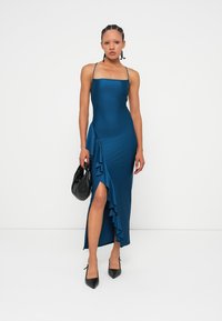 Femme portant une robe midi sans manches en teal avec une fente froncée, tenant un sac à main noir et portant des talons noirs sur un fond blanc uni.