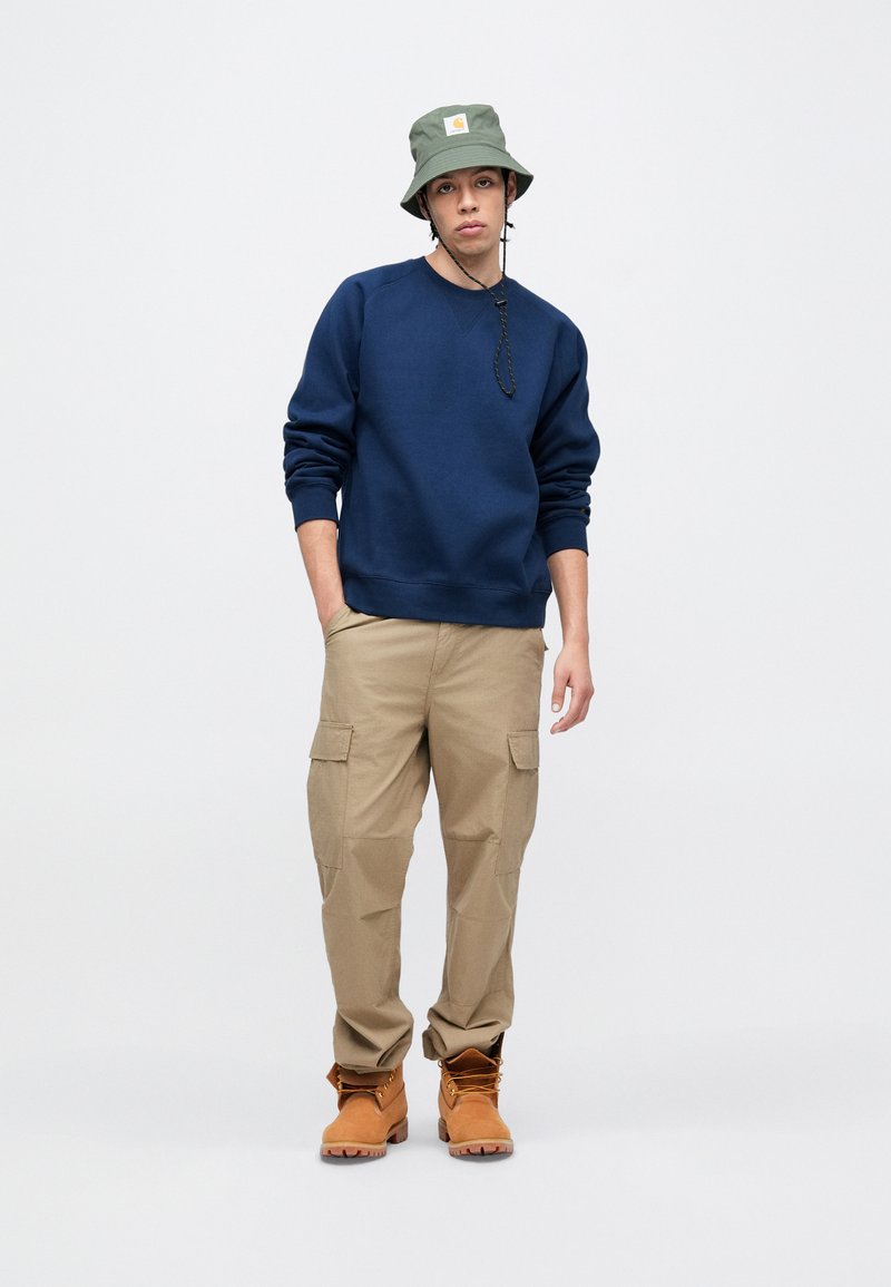 Marineblå sweatshirt, beige cargobukser og brune støvler. Buckethatten er olivengrøn med et hvidt logopatch. Enkel design med funktionelle detaljer.