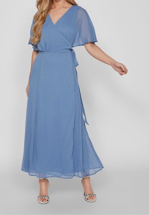 Femme portant une robe portefeuille bleue fluide avec des manches courtes transparentes et des sandales argentées à talons, debout devant un fond uni.